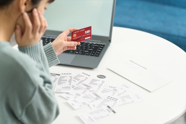 クレジットカードと家計のレシートを見ながら生活費の支払いに頭を抱える主婦。借金やリボ払い、自転車操業の苦しさを象徴